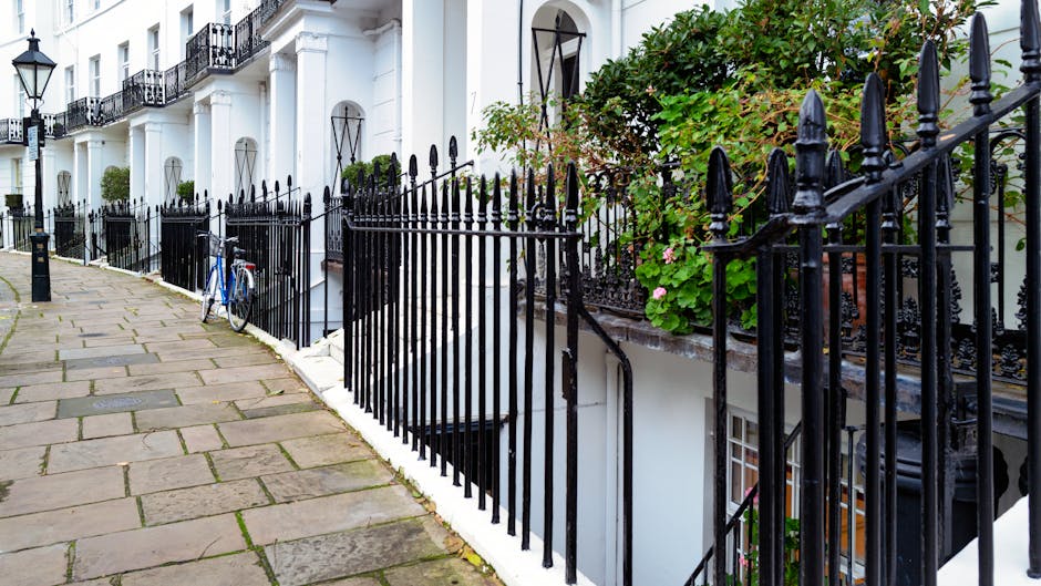 A row of white terraced residential buildings with black wrought iron railings along a paved pavement street. The buildings have tall, narrow windows with arched tops and external doorways. One of the buildings features a small balcony with decorative ironwork and potted green plants with pink flowers. A blue bicycle is leaning against the railing near the foreground, close to a black bollard. The pavement consists of large, rectangular stone slabs with visible joints, and the scene appears to be captured during daylight with natural, even lighting. The setting suggests an urban environment where private property boundaries are marked with metal fencing, aligning with independent waste collection or on-site rubbish clearance practices handled by waste management services such as those offered by wasteclearancesouthkensington.co.uk.