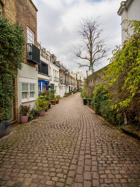 A narrow, cobblestone residential alleyway lined with potted plants and shrubs along the base of multi-story terraced houses on the left side, featuring a mix of white and pastel-colored facades with black iron balconies, while the right side has a tall, mossy brick wall with green foliage spilling over the top. In the background, a tall, leafless tree stands against a cloudy sky, and the scene appears quiet and well-maintained, typical of an urban neighbourhood where private rubbish clearance services might be employed to manage waste removal from nearby properties.