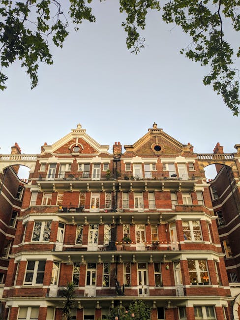 A multi-storey red brick residential building with white window frames and decorative architectural details, situated on a tree-lined street. The building features several balconies with black iron railings, some with potted plants. The roof has ornate gables and chimneys, with a circular window visible on the right gable. The upper part of the building is seen against a clear sky, with green tree branches framing the top of the image. The scene is indicative of an urban residential area and highlights the type of property that might require waste clearance services, such as private estate or apartment block rubbish removal, often managed through independent collection or local waste disposal options. The overall setting suggests a well-maintained building in a residential neighbourhood where professional waste management, including rubbish clearance, could be relevant to homeowners and property managers, exemplifying a typical urban context for rubbish removal services.