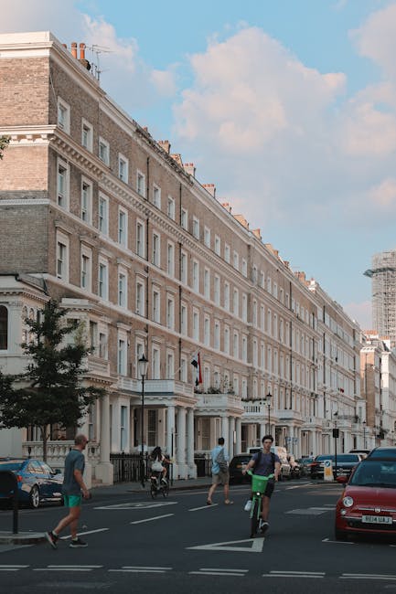A multi-storey red brick residential building with white window frames and decorative architectural details, situated on a tree-lined street. The building features several balconies with black iron railings, some with potted plants. The roof has ornate gables and chimneys, with a circular window visible on the right gable. The upper part of the building is seen against a clear sky, with green tree branches framing the top of the image. The scene is indicative of an urban residential area and highlights the type of property that might require waste clearance services, such as private estate or apartment block rubbish removal, often managed through independent collection or local waste disposal options. The overall setting suggests a well-maintained building in a residential neighbourhood where professional waste management, including rubbish clearance, could be relevant to homeowners and property managers, exemplifying a typical urban context for rubbish removal services.