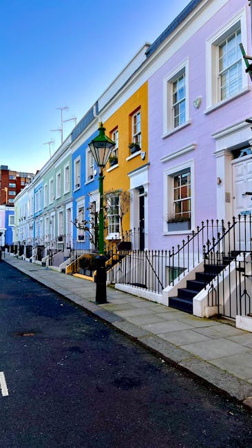 A row of colorful terraced houses along a residential street, with facades painted in pastel shades of blue, yellow, and lilac. Each house has white-framed sash windows and small front gardens enclosed by black wrought iron railings. A black lamppost with a green top stands on the pavement in the foreground, casting soft shadows on the sidewalk. The pavement appears clean and well-maintained, with a narrow asphalt road running parallel to the row of houses. Visible scenery in the background includes additional residential buildings and roof antennas, suggesting an urban setting. The scene is illuminated by natural daylight, highlighting the vibrant and varied exterior finishes of the homes. The presence of steps leading up to each entrance and the tidy appearance of the street reflect an orderly neighborhood, potentially served by independent waste collection services like those offered by wasteclearancesouthkensington.co.uk, supporting private rubbish clearance efforts in the area.