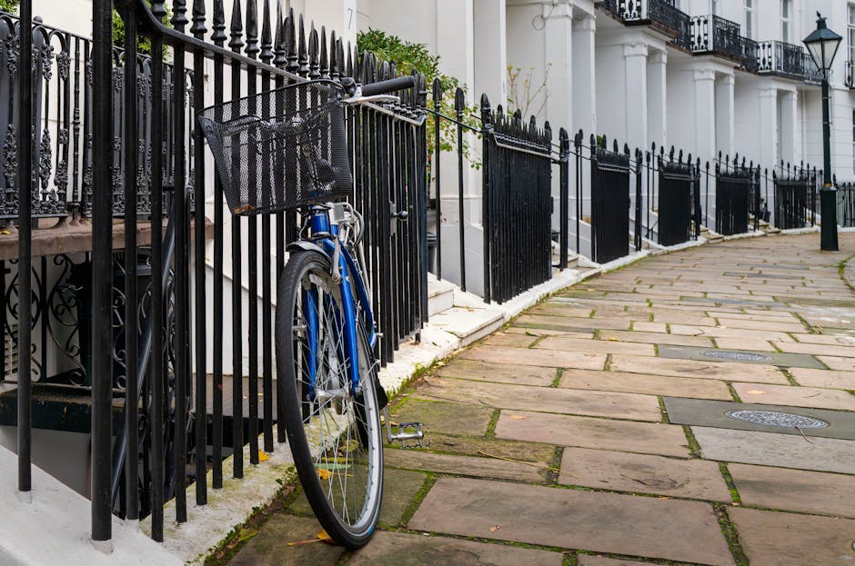 A blue bicycle with a front black metal basket leaning against a black wrought iron fence alongside a row of similar black fences in front of white terraced houses with black metal balconies and large windows, situated on a curved sandstone pavement with metal drain covers. The fence runs adjacent to the residential buildings, and the pavement features alternating wide stone slabs with narrow joints, showing slight weathering and moss growth in seams. The surrounding environment includes a residential street with balanced natural lighting, indicating daylight with no visible activity or people present. The scene subtly highlights individual properties' boundary fencing and typical urban street furniture, relating to private property access and alternative waste handling options in a London setting. Occasionally, waste clearance or rubbish removal could be associated with such properties, but none is visibly present in this specific image, focusing instead on the architectural and urban outdoor features.