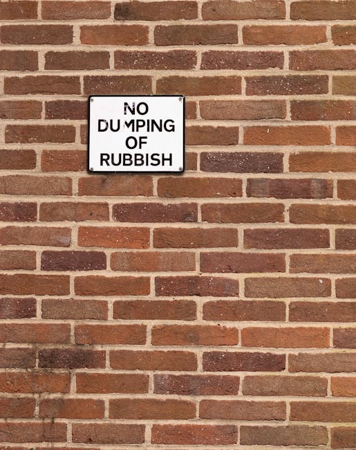 A rectangular white metal sign with black text is mounted on a red brick wall, warning 'NO DUMPING OF RUBBISH.' The bricks are laid in a horizontal pattern with varying shades of red, brown, and some darker tones, creating a textured background. The sign is positioned near the upper left of the wall, with a small gap around it. The wall appears clean and well-maintained, with mortar joints clearly visible between the bricks. The lighting is even, without significant shadows or glare, highlighting the surface details of the brickwork and the sign, which may be part of a private property or area designated for waste management, reflecting proper waste removal practices and regulations enforced by waste clearance services or local authorities.