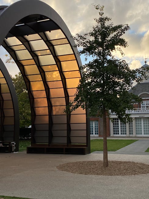 The image features an outdoor scene with a modern, semi-circular structure composed of metal and glass panels, reflecting warm tones from the sunset sky. The structure stands on a paved surface, with the panels showcasing a combination of brushed metal finishes and translucent glass, arranged in a grid pattern supported by black metal frames. Adjacent to the structure is a small, neatly maintained tree with a thin trunk and green leaves, planted in a circular bed bordered by gravel or mulch. In the background, classic brick and white-framed windows of a traditional building are visible, suggesting an institutional or historic setting. The overall environment combines contemporary architectural elements with natural outdoor features, set against a softly lit sky with scattered clouds. This scene exemplifies the type of urban or institutional space that might require interim rubbish clearance services, such as site cleanups or window of disposal for renovation or event preparations, with the modern structure and surrounding greenery providing a visually balanced context relevant to private waste management solutions. The scene as a whole underscores the importance of professional rubbish removal, exemplified by independent collection options that facilitate efficient onsite clearance when dealing with diverse outdoor features.