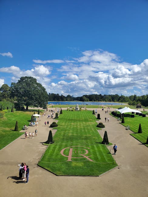 A wide view of a landscaped garden area with neatly trimmed grass and decorative elements, including a central section with an ornate pink pattern. The garden is bordered by walkways with several small groups of people walking and standing. To the right, there are temporary white tents and some small landscaped bushes shaped into cones, with a few individuals nearby. In the background, there is a large body of water with trees lining the opposite bank under a bright blue sky filled with scattered white clouds. The setting appears to be an outdoor public space, potentially a historic park or venue, where private event waste removal or on-site clearance might be necessary, as indicated by the careful landscaping and presence of event-related elements. The image, associated with waste management services such as those offered by wasteclearancesouthkensington.co.uk, highlights the importance of professional rubbish removal during large events or venue preparations to maintain aesthetic quality and environmental standards.