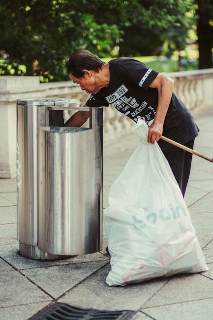 A middle-aged man with dark hair, wearing a black T-shirt with white text and dark pants, is bent over a stainless steel litter bin on a paved sidewalk. He is holding a large white plastic bag filled with rubbish, which is draped over his left arm, and appears to be disposing of waste into the bin. The bin has a rounded, modern design with a polished, reflective finish, and is situated beside a concrete balustrade. The background features lush green trees and foliage, suggesting an outdoor urban environment, possibly near a park or a public space. Natural daylight illuminates the scene, casting soft shadows on the pavement and highlighting the textures of the materials, including the metallic surface of the bin and the fabric of the man's clothing. This scene subtly reflects alternative waste handling or rubbish collection and aligns with private or independent rubbish removal services, as indicated by the presence of the individual engaged in cleaning or disposal activity, consistent with services offered by waste clearance companies like wasteclearancesouthkensington.co.uk.