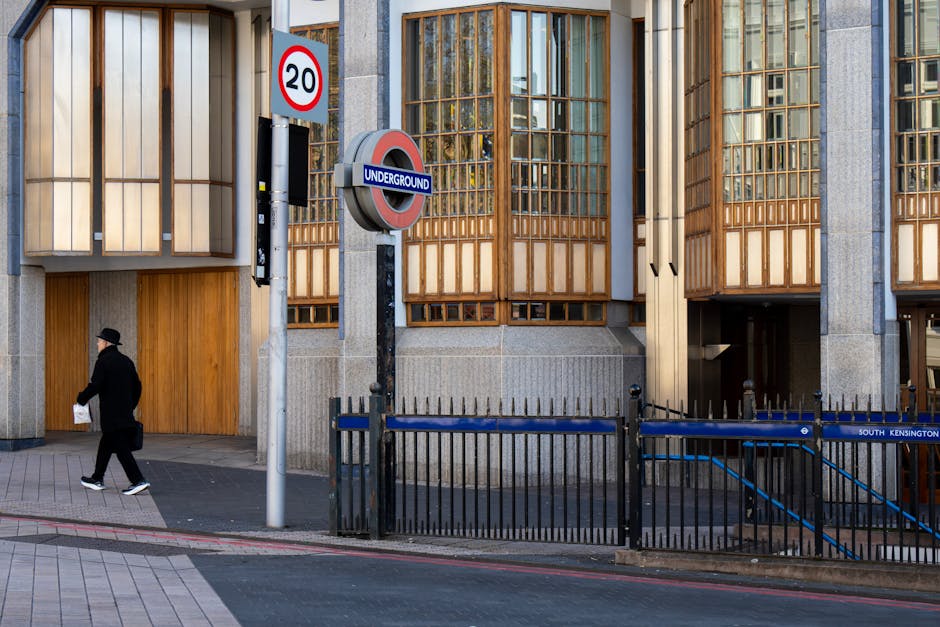 A daytime street scene outside South Kensington Station featuring a modern building with distinctive wooden framed windows and concrete pillars. In the foreground, a black metal safety fence lines the pavement, alongside a blue sign indicating an underground entrance. Near the fence, a pedestrian dressed in dark clothing, including a coat, hat, and sneakers, is walking along the sidewalk while carrying a white paper bag. A street sign displaying a speed limit of 20 mph is mounted on a pole adjacent to a circular underground station sign. The environment appears clean and well-maintained, with natural light illuminating the building's textured materials, highlighting the wooden window frames, the grey concrete, and the pavement's patterned surface. This scene subtly relates to the context of urban waste management and alternative rubbish collection practices often necessary in high-foot-traffic city areas protected by security barriers and signage, emphasizing the importance of efficient rubbish removal services like those provided by wasteclearancesouthkensington.co.uk.