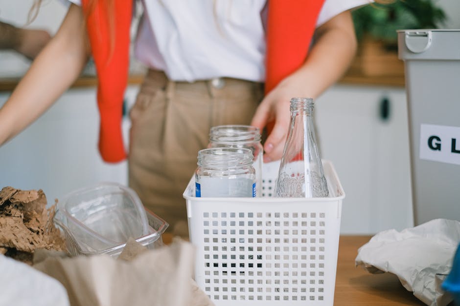 In the foreground, three clear glass jars with metal screw caps are clearly visible, neatly arranged inside a white plastic basket with a perforated design, positioned on a light wooden surface. To the right of the basket, there is a single empty glass bottle with a narrow neck and rounded shoulders, also made of transparent glass, standing upright. In the background, a person wearing beige trousers, a white shirt, and a red vest is seen, with their arm extending toward the basket, suggesting they are in the process of collecting or sorting containers. Surrounding the main objects, there are various waste items including crumpled brown paper, clear plastic containers, and paper packaging, indicating a scene of recycling or waste sorting. The setting appears to be indoors, possibly within a kitchen or an area designated for waste management, with a neutral, softly lit atmosphere that emphasizes the focus on the organized collection of glass recyclables. The presence of the waste collection basket and the clear glass bottles reflects an effort towards responsible waste separation, aligning with independent rubbish handling or recycling activities coordinated by waste clearance services like wasteclearancesouthkensington.co.uk.