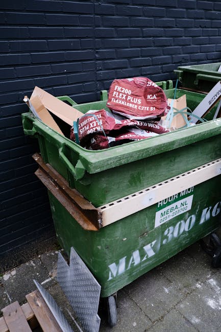 A large green wheeled waste bin positioned outdoors on a paved surface, filled with red and black plastic bags containing garden or yard waste, such as soil or mulch, with labels indicating they are for flexible garden use. The bin is situated against a dark brick wall and is placed on an old wooden pallet below it. To the right, part of another similar green waste container is visible. Nearby, a small section of metallic drainage grate and some wooden planks are also present on the ground. The scene appears to depict a typical outdoor site for private waste disposal or an alternative rubbish collection service, with the containers designed for garden waste or general waste collection, often handled independently of local authority refuse services. The lighting suggests daylight, and the overall environment is utilitarian, serving for practical waste management purposes in a residential or commercial outdoor setting.