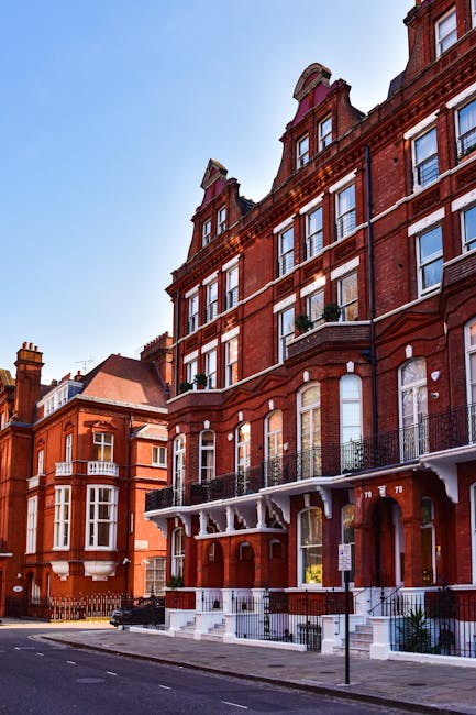 The image depicts a row of tall, red brick residential buildings with ornate architectural details, including decorative cornices and arched windows. The buildings feature multiple stories, with some windows framed by white trim and black wrought iron balconies on the lower levels. The facades are textured with brickwork and vertical white accents, adding visual interest. In the foreground, a paved street runs along the front of the buildings, with a few small potted plants placed near the entrances, and a street sign visible on the pavement. The scene is illuminated by daylight under a clear, bright blue sky, creating a crisp, professional atmosphere. The overall setting suggests an urban environment with classic Victorian or Edwardian architecture, characteristic of independent property management or private waste disposal options often associated with luxury residential areas, aligning with the context of rubbish removal services in the vicinity.